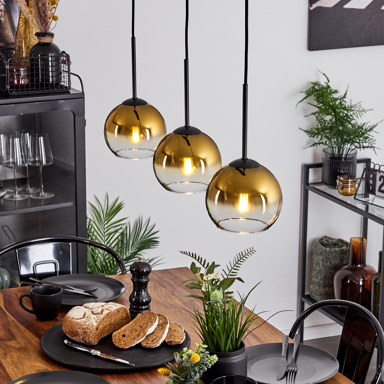 Esstisch mit Brot, Tellern und Hängelampen. Drei goldfarbene Pendelleuchten über einem Holztisch.
