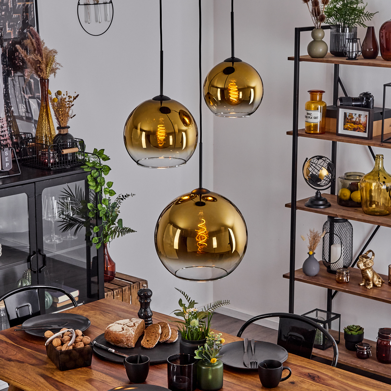 Esszimmer mit Holztisch, goldenen Pendelleuchten und Regal mit Dekoration. Teller, Brot und Pflanzen sind auf dem Tisch zu sehen.