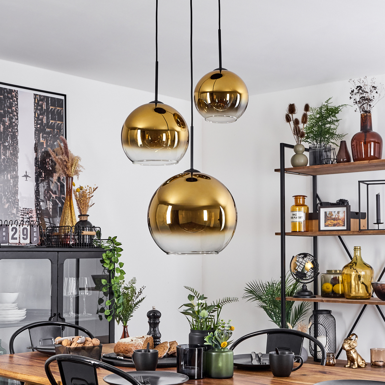 Drei goldene Pendelleuchten hängen über einem Holzesstisch, der für eine Mahlzeit gedeckt ist. Regale mit Dekor befinden sich hinter dem Tisch.