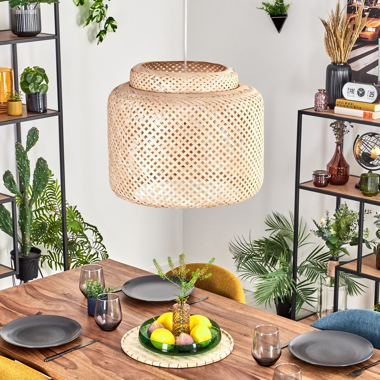 Ein Esstisch mit Tellern, Gläsern und einem Mittelstück aus Zitronen und Feigen. Eine gewebte Lampe hängt darüber. Regale mit Pflanzen sind sichtbar.