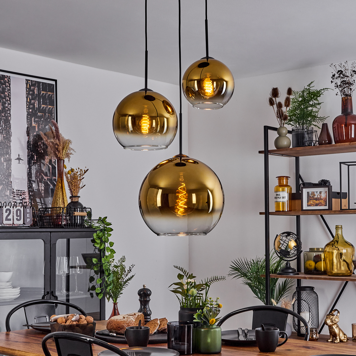 Esszimmer mit goldenen Hängelampen. Ein Holztisch ist mit Essen und Geschirr gedeckt. Regale im Hintergrund halten Deko.