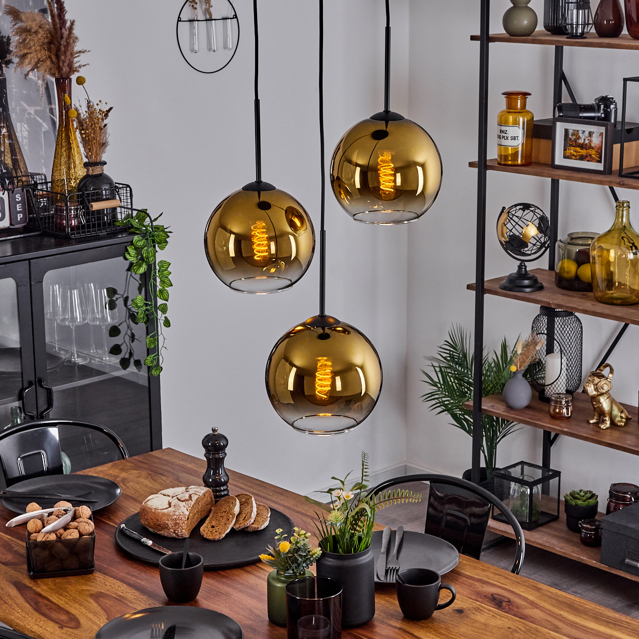 Esszimmer mit Holztisch, drei Hängelampen, Brot, Tellern und einem Regal mit Dekorationen.
