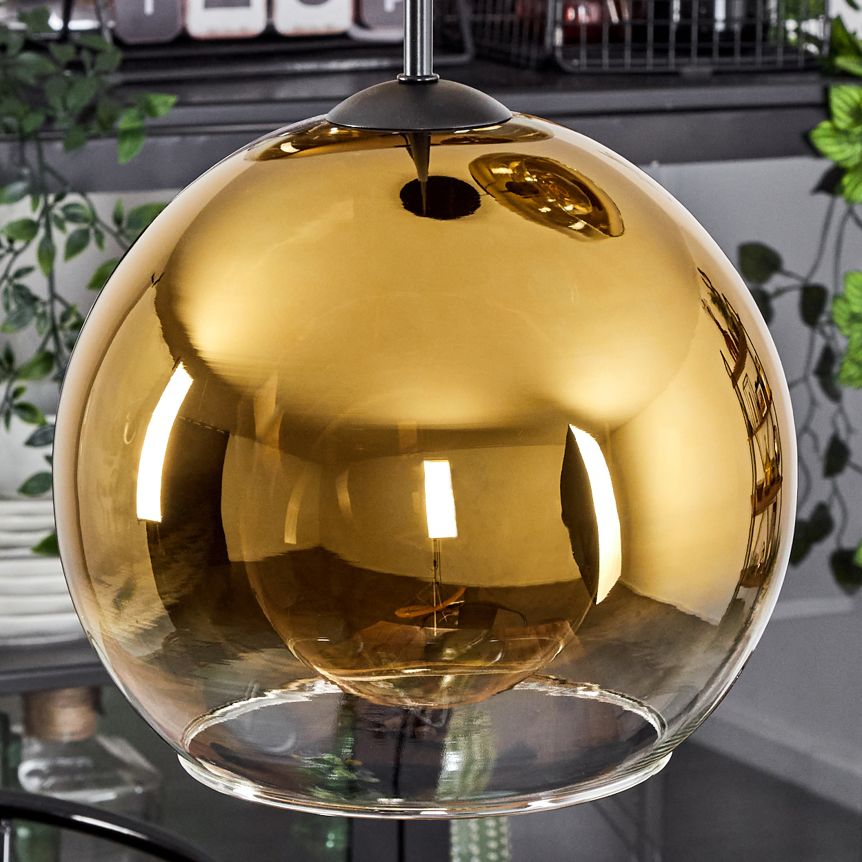 Eine goldene, kugelförmige Pendelleuchte mit sichtbarer Glühbirne im Inneren, die an einer Metallstange hängt. Der Hintergrund ist eine verschwommene Küchenszene.