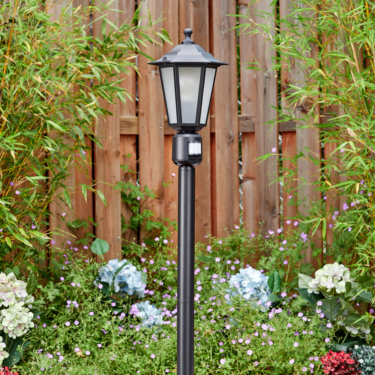 Schwarze Außenlampe im Garten. Bambus und Holzzaun im Hintergrund. Blumen im Vordergrund.