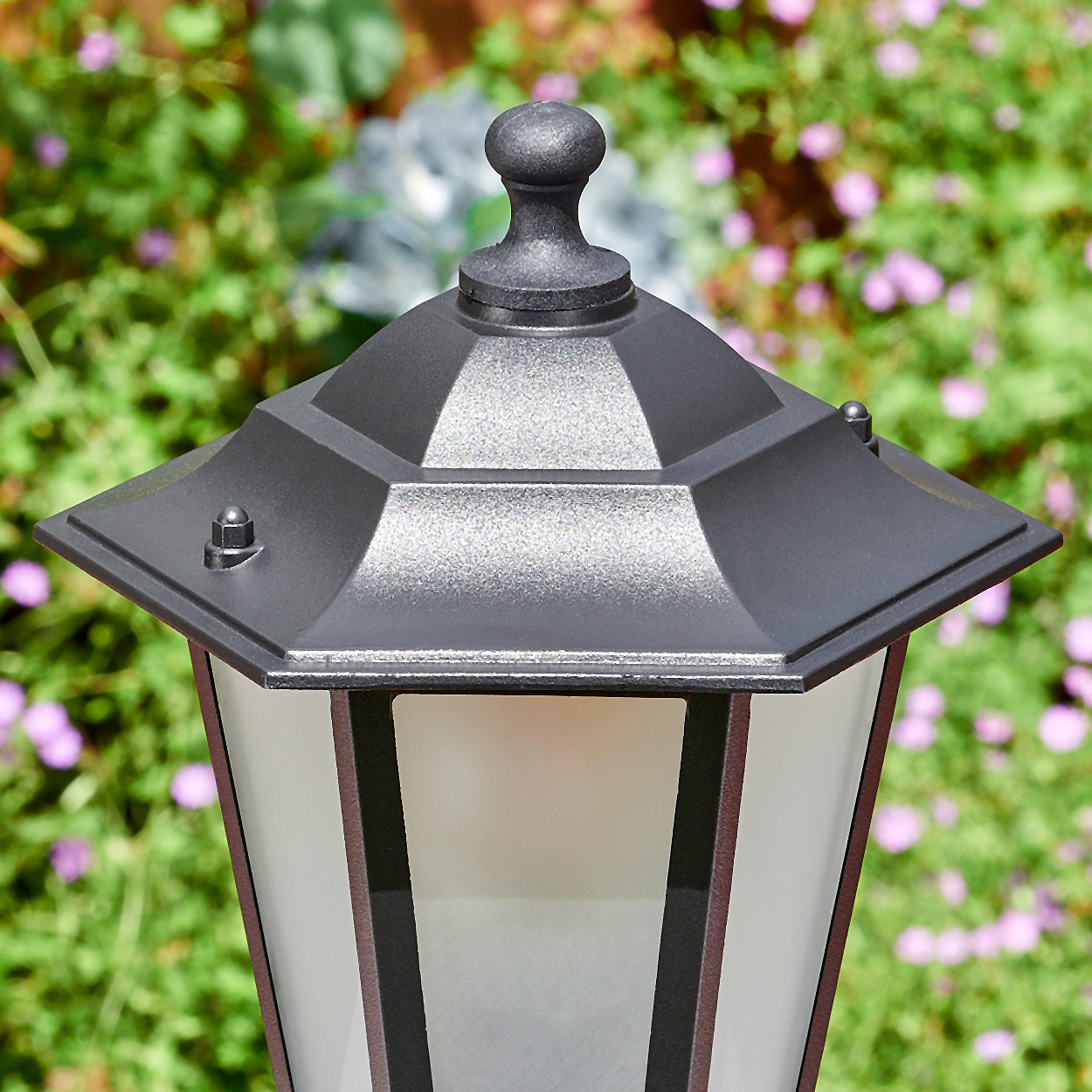 Nahaufnahme einer schwarzen Außenlampe mit sechseckiger Oberseite und Milchglasscheiben, vor verschwommenem grün-lila Blumengrund.