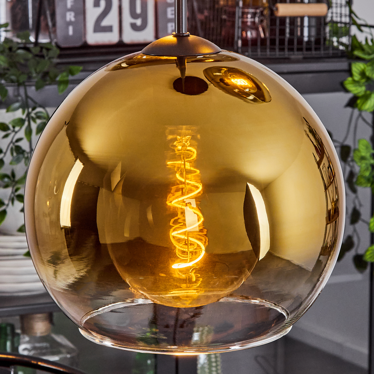 Eine goldene Glaskugel-Pendelleuchte mit einer Wendelglühbirne. Im Hintergrund sind ein Kalender und etwas Grün zu sehen.
