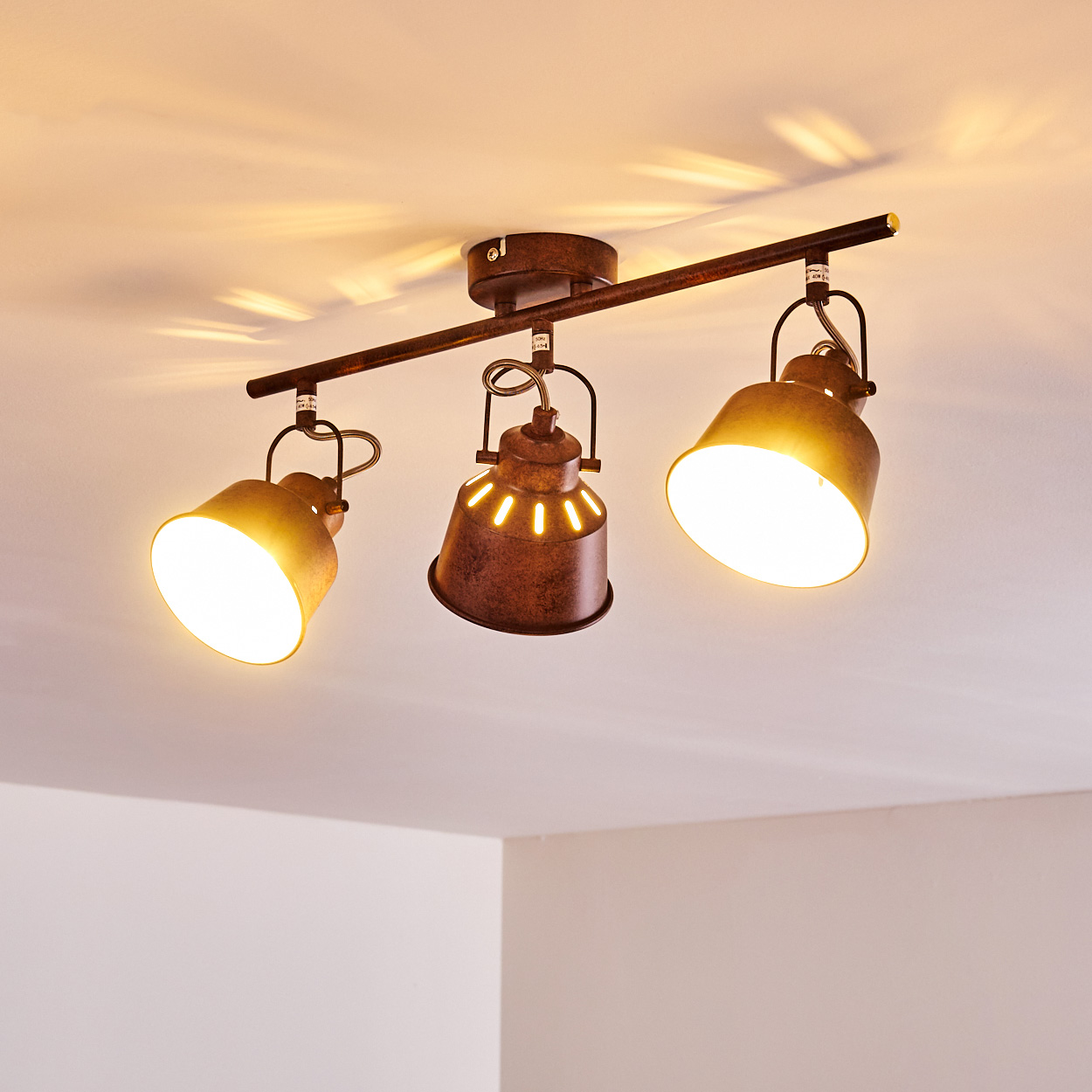 Deckenstrahler mit drei Lichtern. Die Lampen sind braun und leuchten. Der Hintergrund ist eine weiße Decke und Ecke.