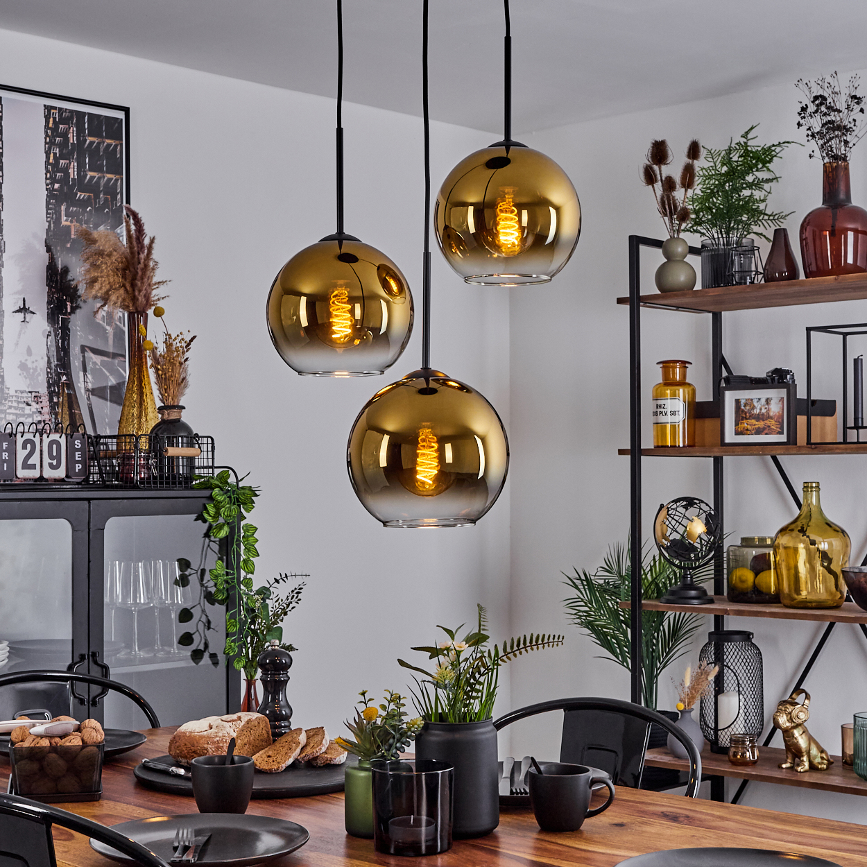Esszimmer. Drei hängende Kugelleuchten. Tisch mit Tellern, Brot und Blumen. Regale mit Dekoration im Hintergrund.