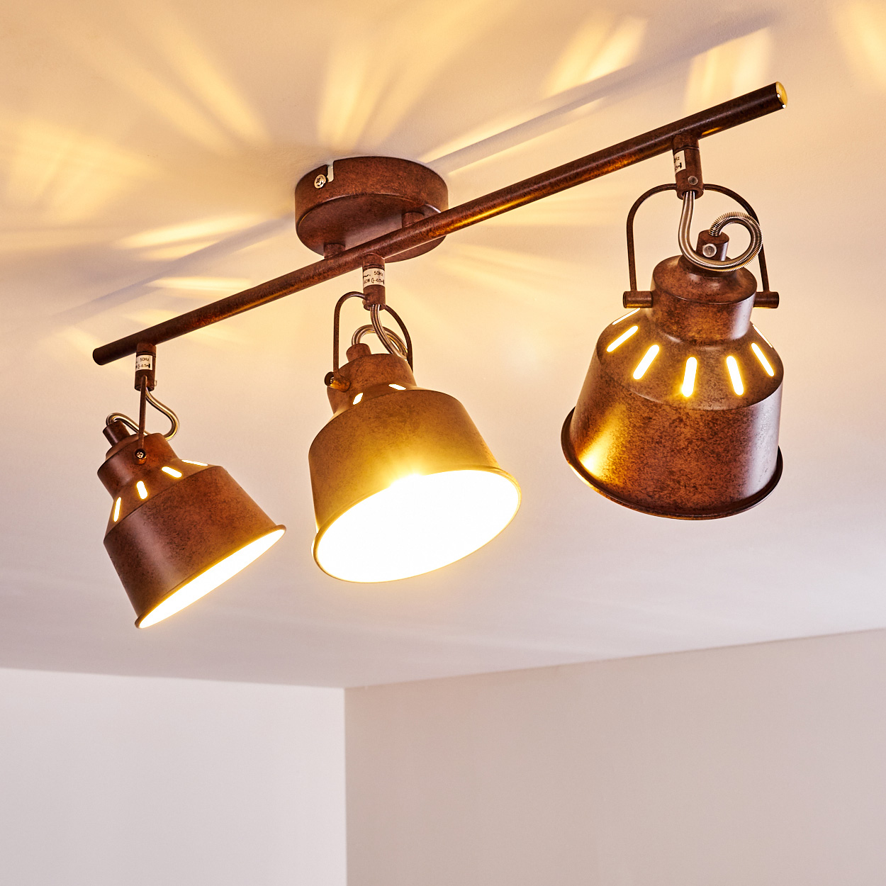 Drei rostbraune Strahler an einer Stange, an einer weißen Decke befestigt. Die Lichter sind an und beleuchten den Raum.