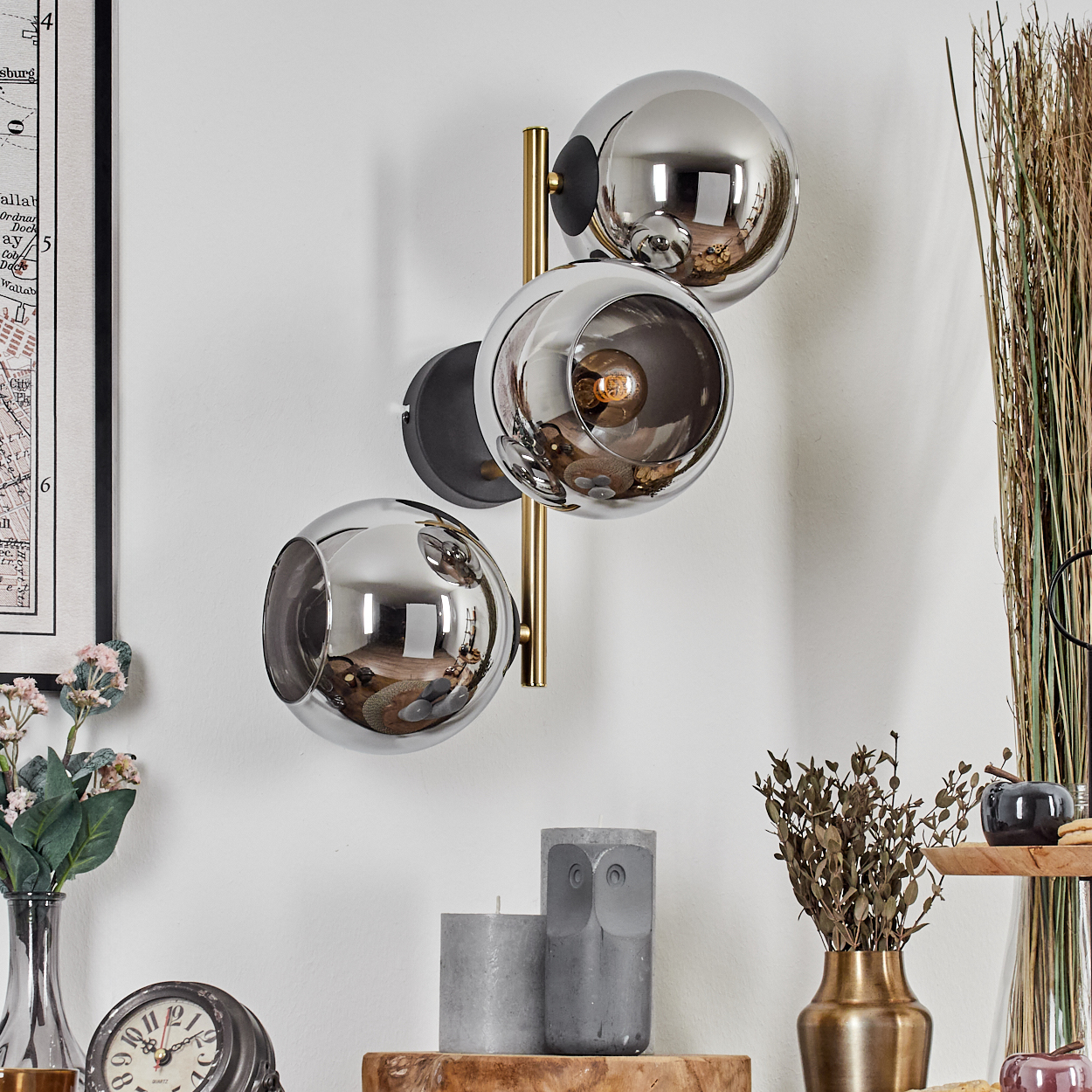 Eine Wandlampe mit drei kugelförmigen Schirmen. Die Raumdekoration umfasst Kerzen, Vasen und eine Uhr.