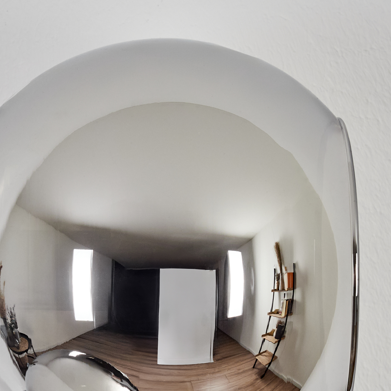 Eine reflektierende Oberfläche, die einen Raum zeigt. Der Raum hat einen Holzboden, weiße Wände und ein Leiterregal mit Büchern.