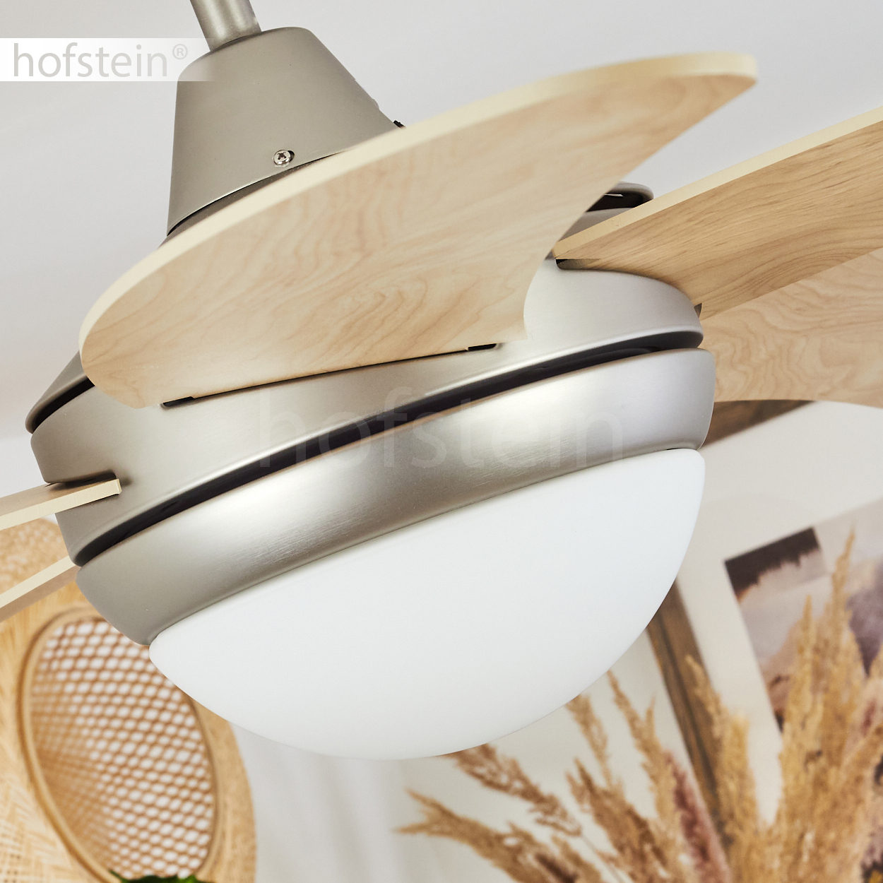 Nahaufnahme eines Deckenventilators mit Holzflügeln und einem weißen Licht. Der Lüfter ist silberfarben. Der Hintergrund beinhaltet Dekoration.