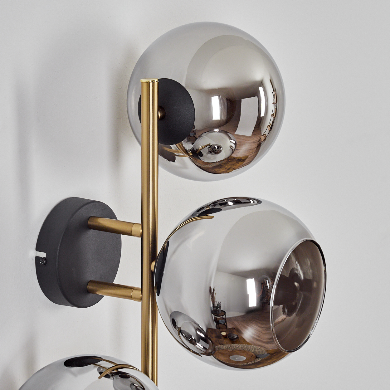Moderne Wandlampe mit drei kugelförmigen, verspiegelten Glasschirmen, getragen von goldfarbenen Metallarmen an einer weißen Wand.