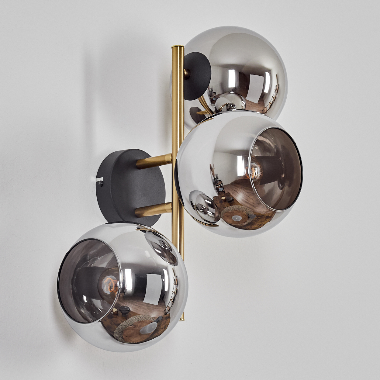 Eine moderne Wandlampe mit drei reflektierenden, kugelförmigen Schirmen. Die Leuchte hat goldfarbene Arme und einen schwarzen Sockel, montiert an einer weißen Wand.