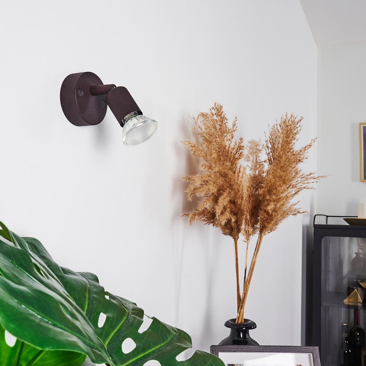 Eine braune Wandleuchte an einer weißen Wand. Getrocknete Pflanzen in einer Vase. Ein grünes Blatt im Vordergrund.