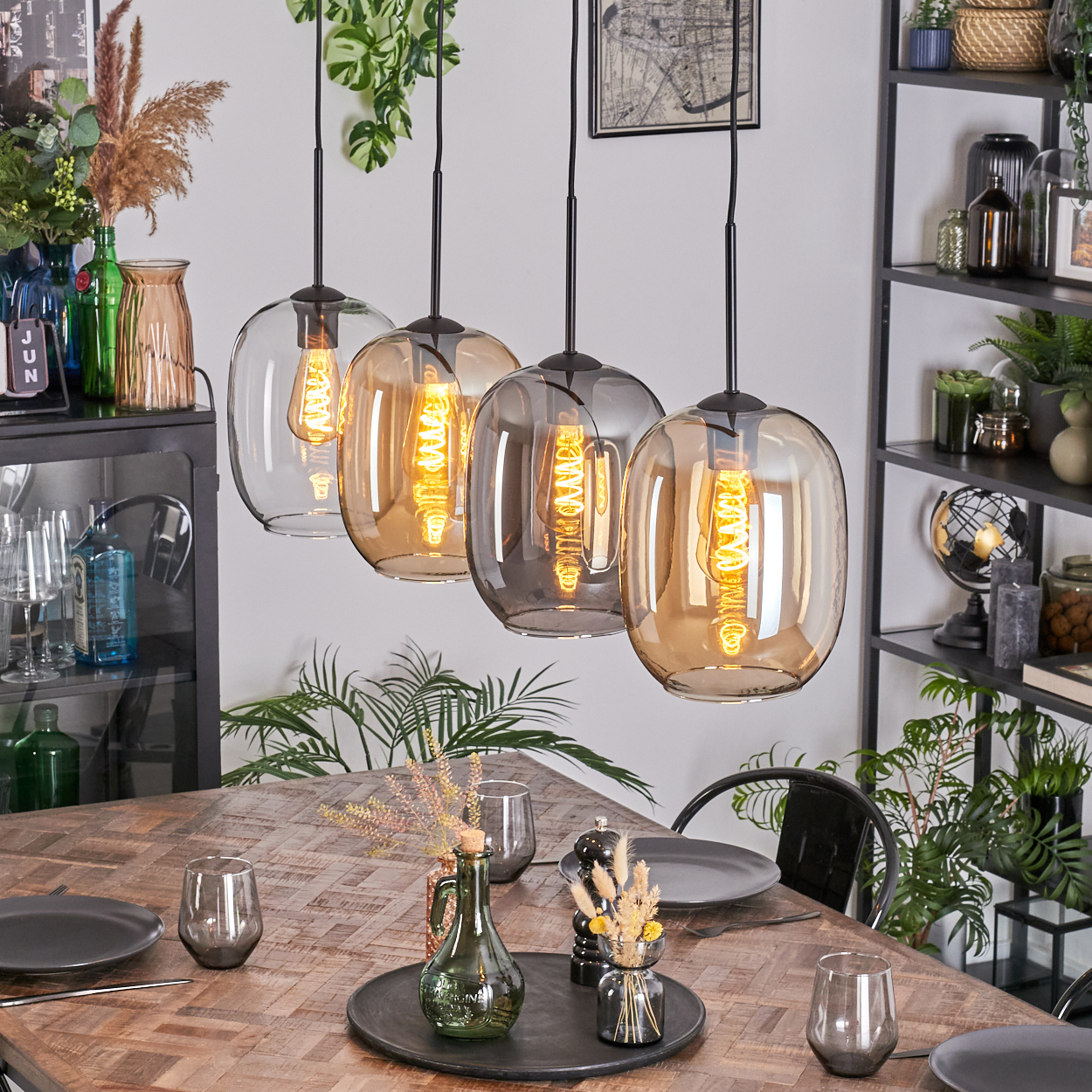 Esszimmer mit Holztisch, Hängelampen und Dekoration. Enthält Teller, Gläser und eine dekorative Vase. Regale mit Gegenständen im Hintergrund.