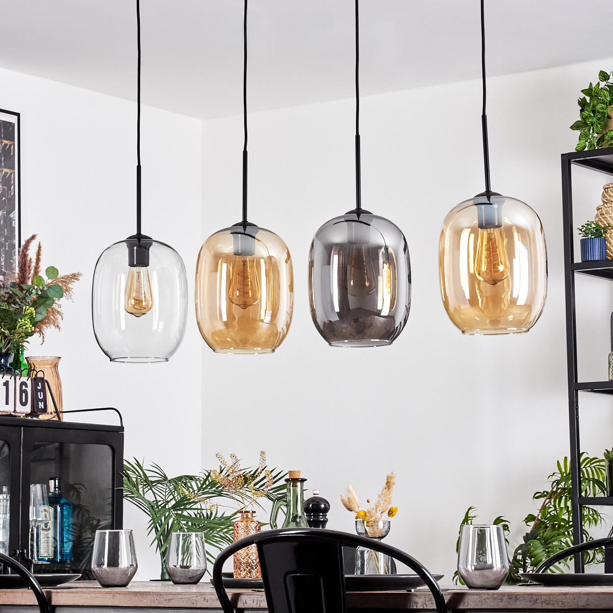 Vier Pendelleuchten mit Glasschirmen hängen über einem Esstisch. Die Szene umfasst Pflanzen, Gläser und ein schwarzes Metallregal.