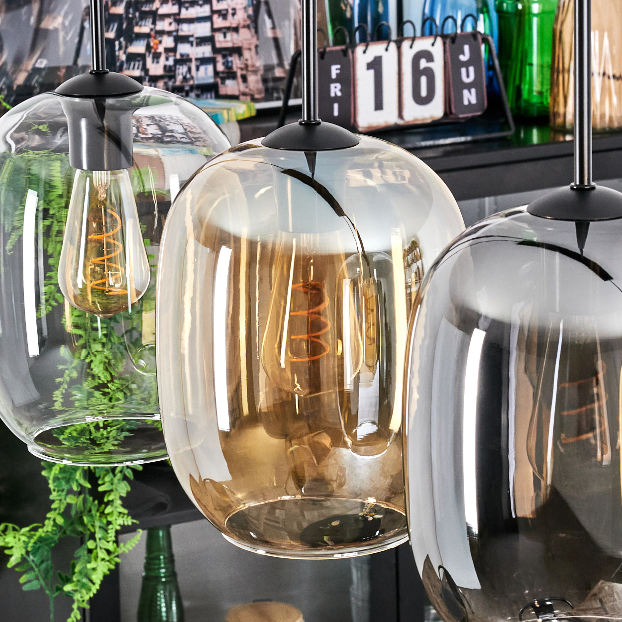 Drei Glas-Hängelampen mit sichtbaren Glühbirnen. Im Hintergrund ein Kalender, grüne Pflanzen und Glasflaschen.
