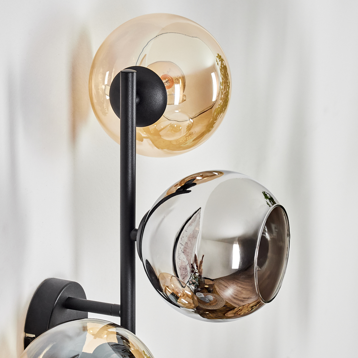 Eine moderne Wandlampe mit drei kugelförmigen Schirmen: bernsteinfarben, verspiegelt und nochmals bernsteinfarben. Ein schwarzer Metallrahmen.