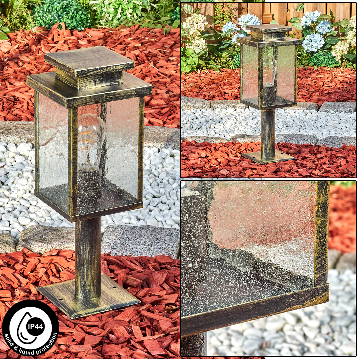 Eine bronzefarbene Außenlampe mit Glasgehäuse. Sie steht auf einer Basis aus roter Mulche und weißen Steinen, mit Pflanzen im Hintergrund.