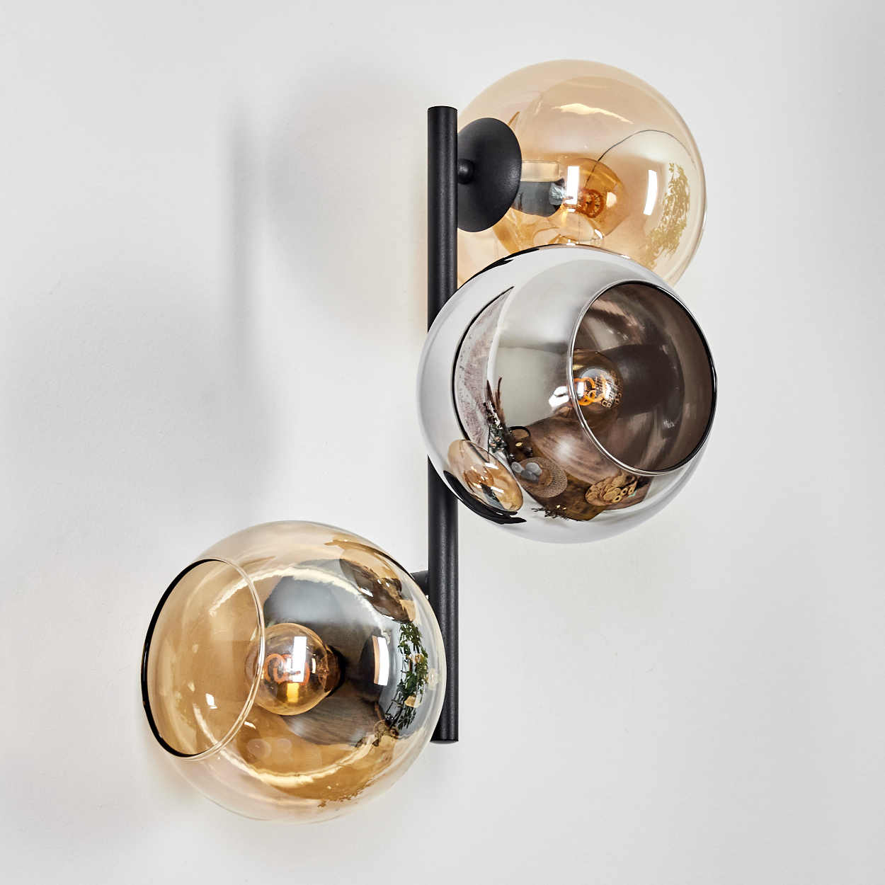Eine moderne Wandlampe mit drei kugelförmigen Schirmen in Gold und Silber, montiert auf einem schwarzen Metallrahmen, vor einer weißen Wand.