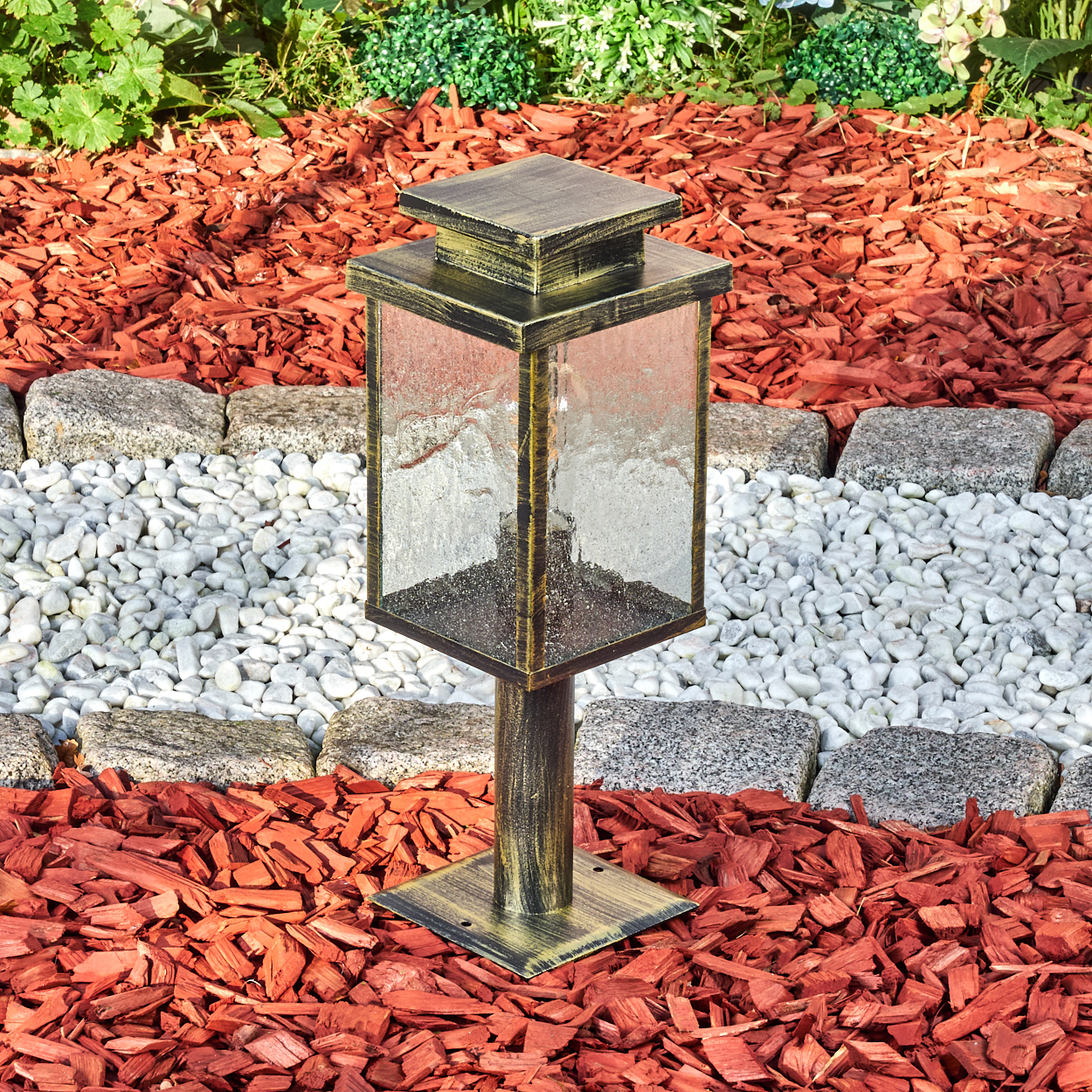 Eine dekorative Lampe steht auf roter Mulch, mit einem Hintergrund aus weißen Kieselsteinen und grünen Pflanzen. Die Lampe ist quadratisch, mit Glasscheiben.