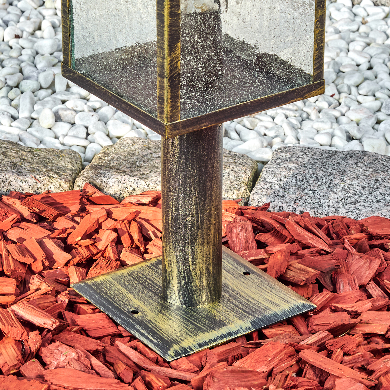 Eine bronzefarbene Außenlampe steht auf roter Mulch. Im Hintergrund befinden sich weiße Kieselsteine und graue Steine.