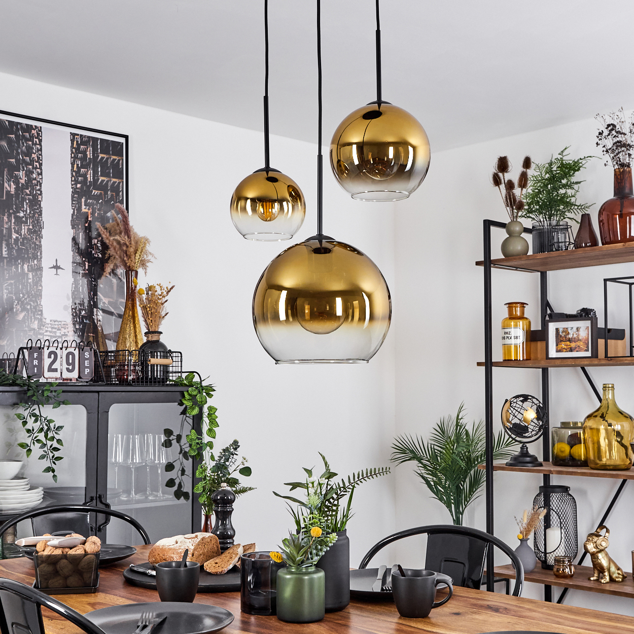 Esszimmer mit gedecktem Tisch. Drei goldene Pendelleuchten hängen darüber. Regale und ein Schrank sind sichtbar.