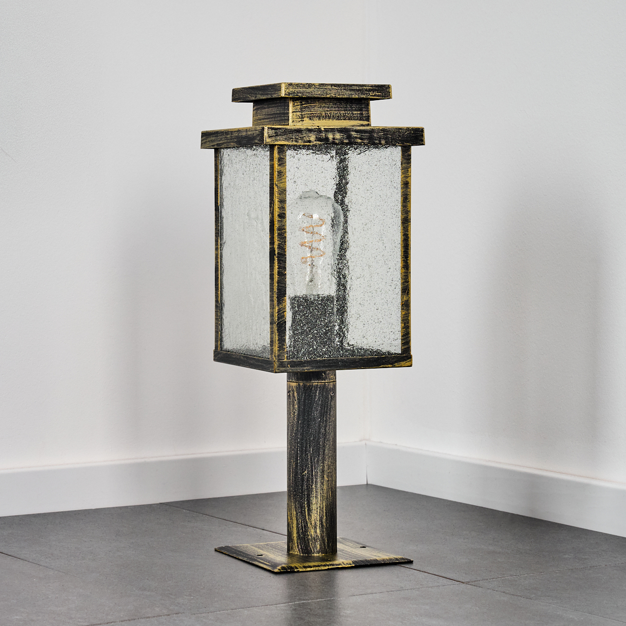 Eine dekorative Außenlampe mit quadratischem Glaskasten, goldenen Akzenten und einer sichtbaren Glühbirne. Sie steht auf einem gefliesten Boden.