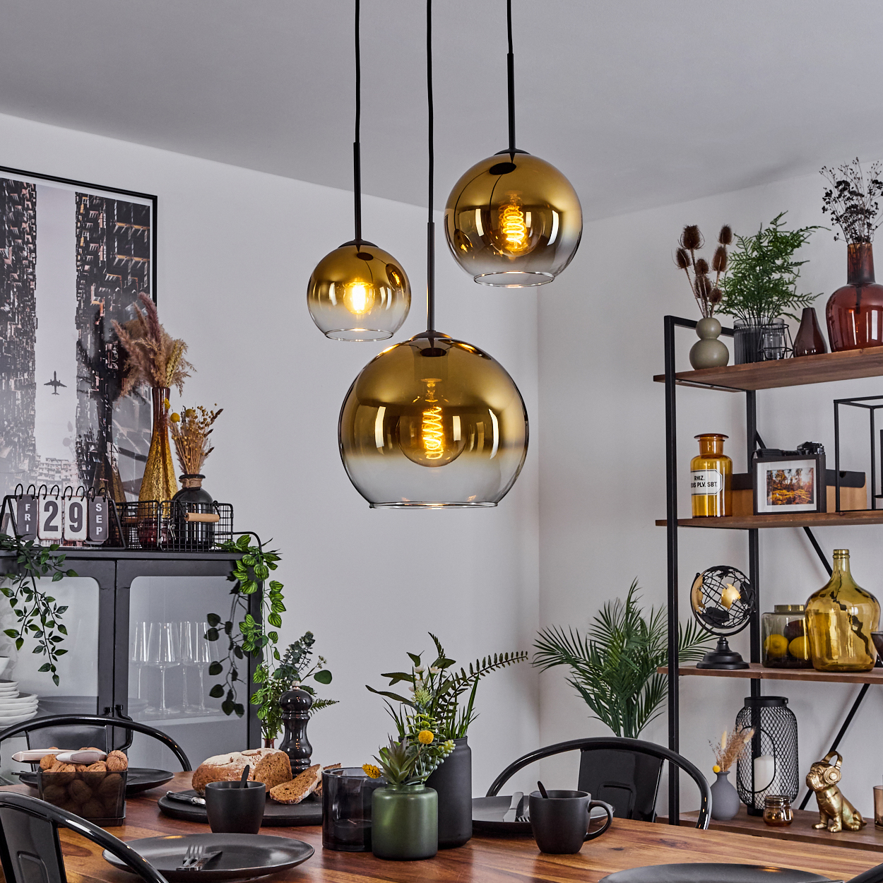 Esszimmer mit golden getönten Hängelampen. Tisch mit Tellern, Brot und Pflanzen gedeckt. Regale mit Dekoration sind im Hintergrund sichtbar.