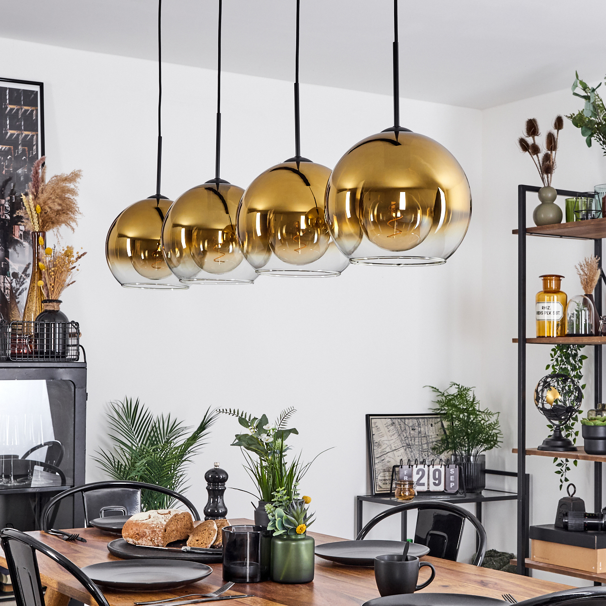 Esszimmer mit Holztisch, gedeckt für das Abendessen. Goldene Hängeleuchten hängen darüber. Regale mit Deko rechts.