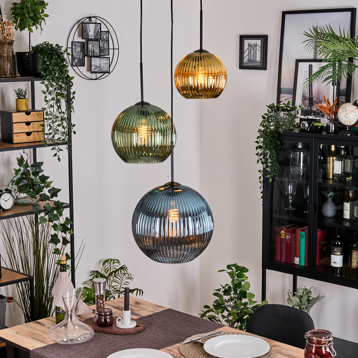 Esszimmer mit drei Hängelampen. Pflanzen und Dekoration umgeben einen Holztisch, der mit einem Glasschrank zum Abendessen gedeckt ist.