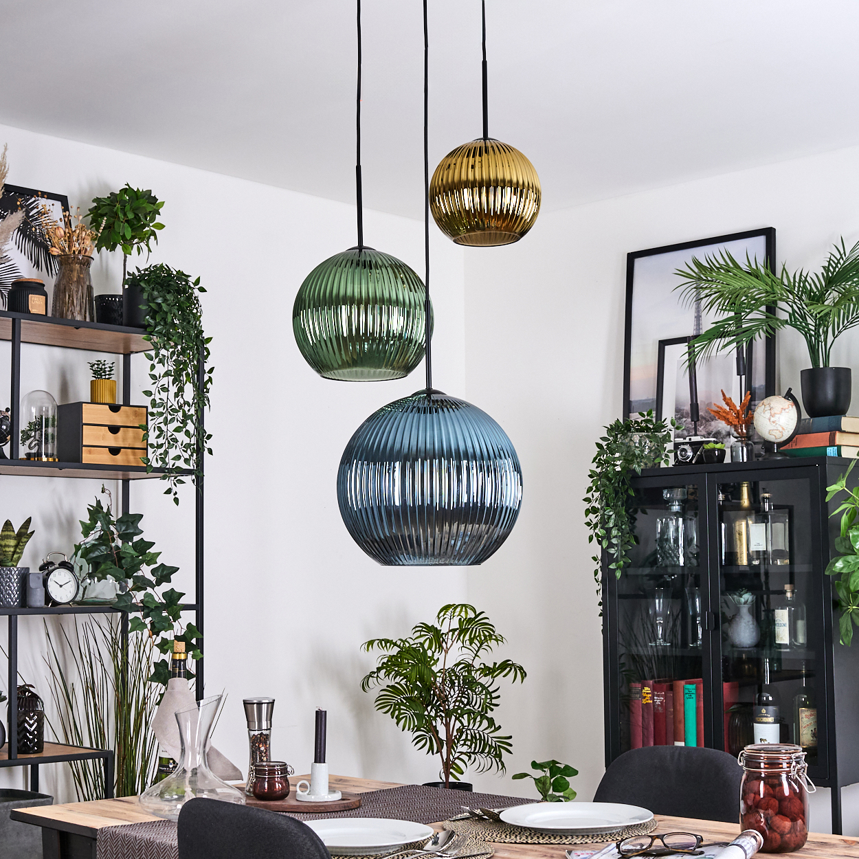 Drei Pendelleuchten über einem Esstisch. Die Leuchten sind grün, blau und goldfarben. Pflanzen und ein Schrank sind im Hintergrund.