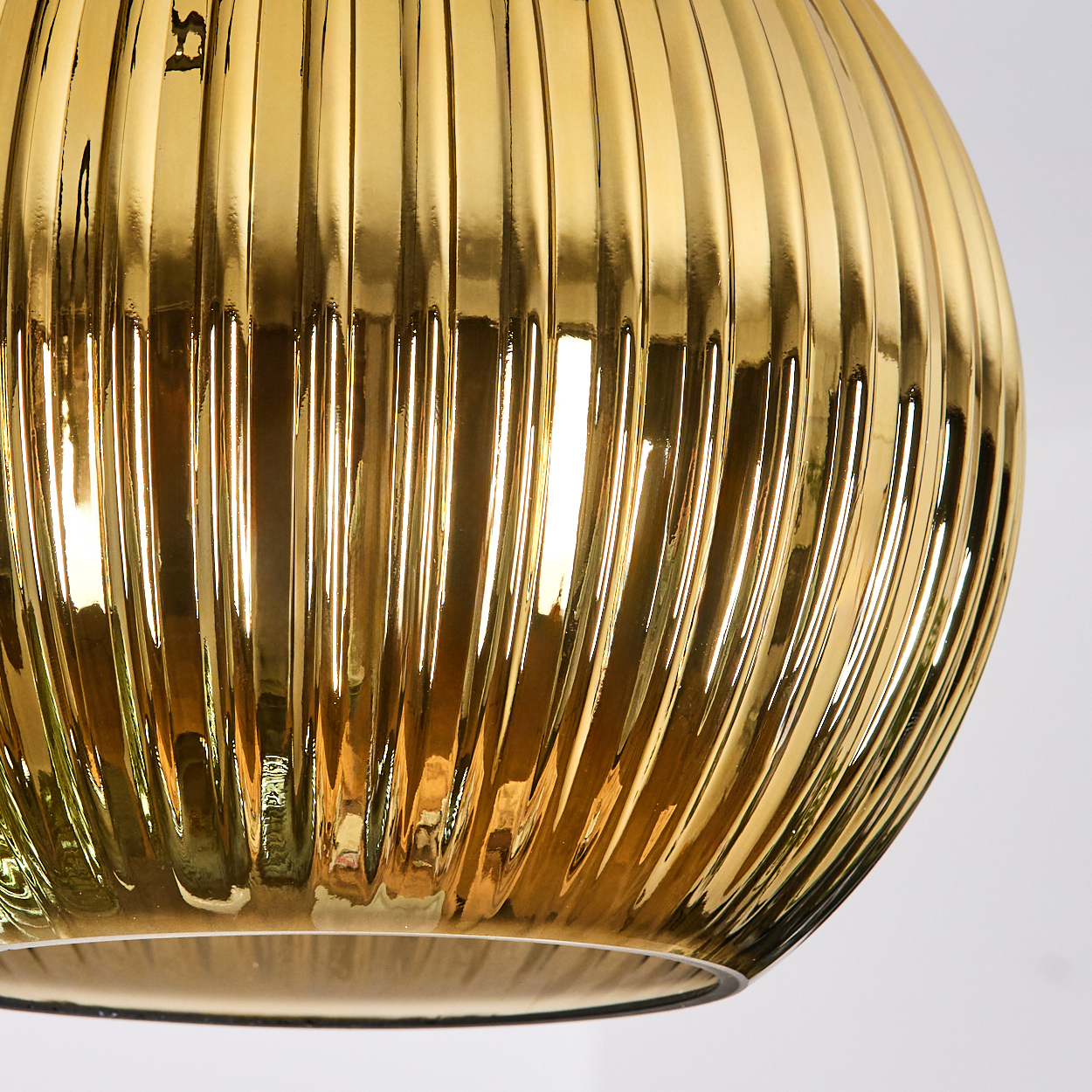 Nahaufnahme eines goldenen, gerippten Glasschirmes. Licht reflektiert auf der Oberfläche und erzeugt Glanzlichter und Schatten. Der Hintergrund ist weiß.