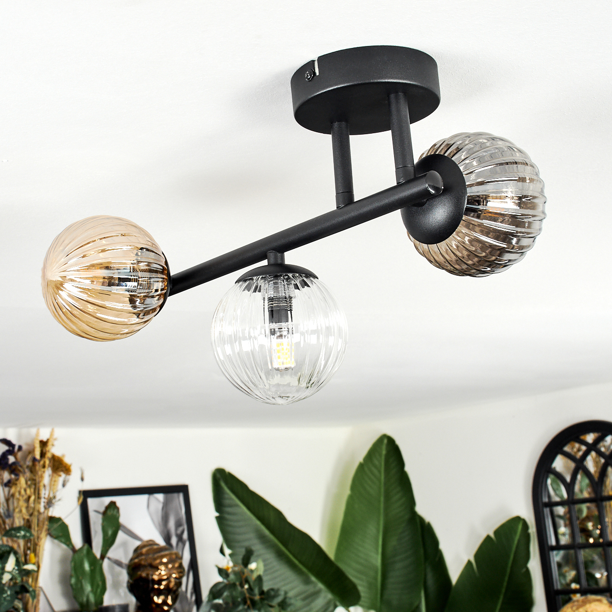 Eine moderne Deckenlampe mit drei Glaskugeln in Bernstein und Grau, montiert auf einem schwarzen Metallrahmen, mit Pflanzen im Hintergrund.