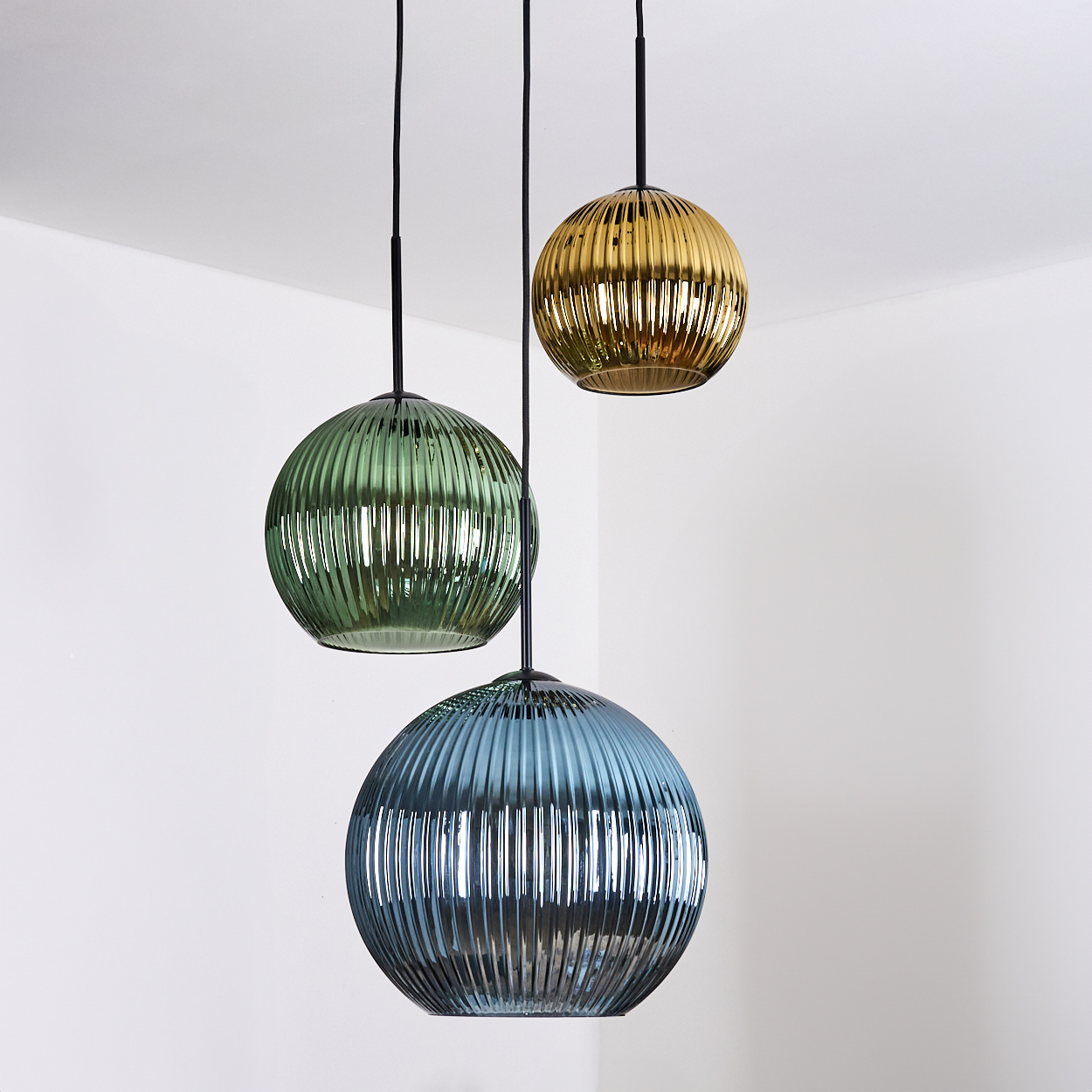 Drei kugelförmige Pendelleuchten, grün, gold und blau, hängen vor einer weißen Ecke. Jede hat eine geriffelte Oberfläche.