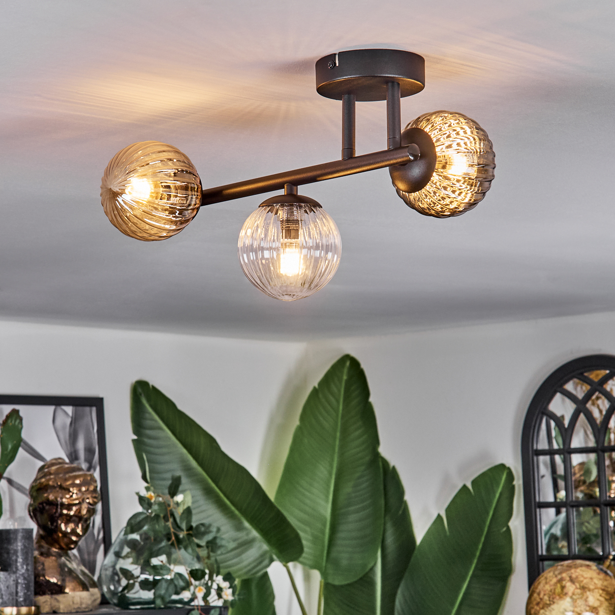 Deckenlampe mit drei Glaskugeln. Grüne Pflanzen und eine Büste im Vordergrund. Der Hintergrund ist eine weiße Decke.