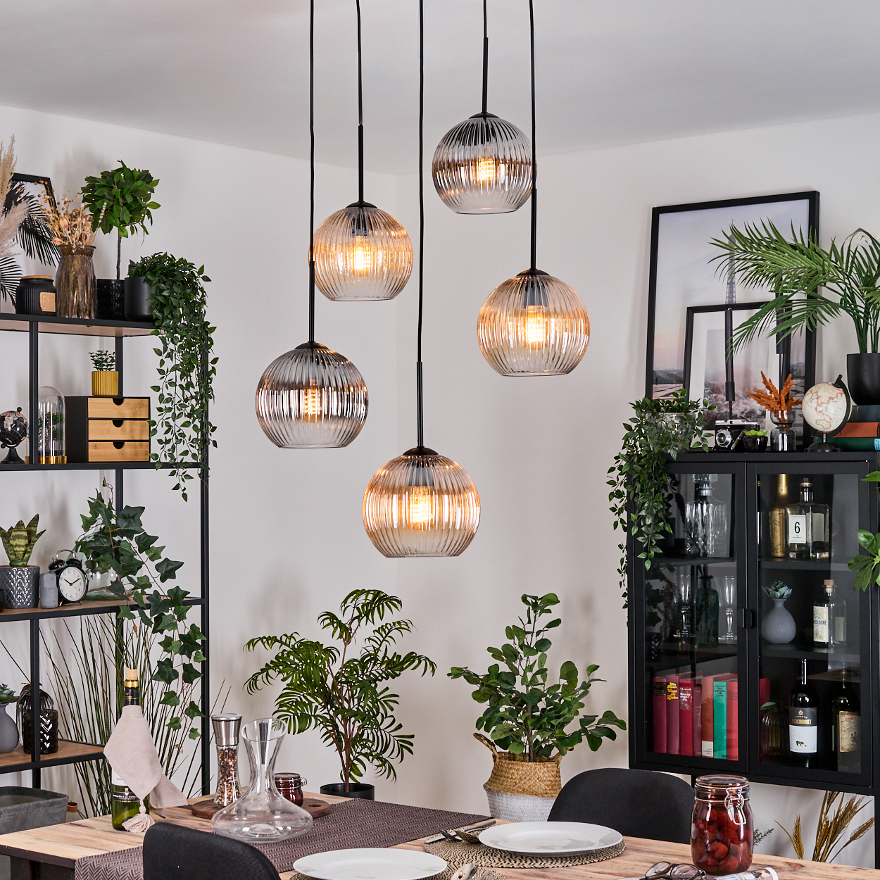 Esszimmer mit Hängelampen, Pflanzen und einem gedeckten Tisch. Schwarze Regale mit Dekor und ein Schrank.