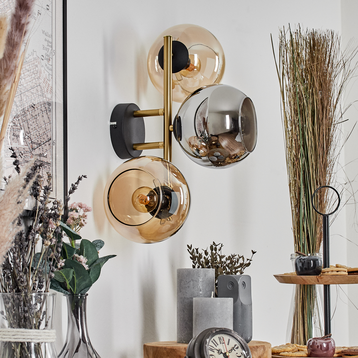 Eine Wandlampe mit drei Glaskugeln. Darunter verschiedene Dekorationsartikel, darunter Kerzen, Blumen und eine Uhr.