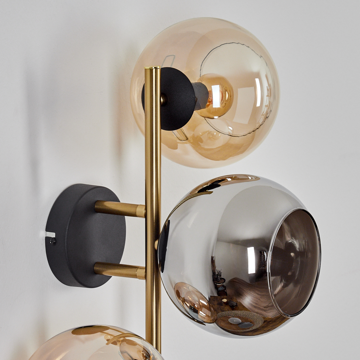 Eine moderne Wandlampe mit drei kugelförmigen Schirmen in Gold und Silber, montiert auf einem goldenen Rahmen, an einer weißen Wand.