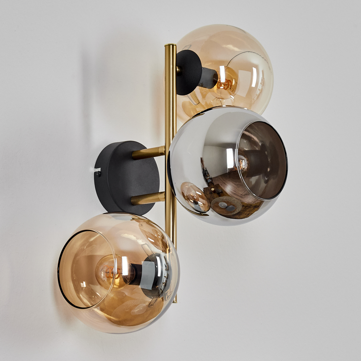 Eine moderne Wandlampe mit drei kugelförmigen Schirmen. Zwei bernsteinfarbene Glaskugeln und eine silberne, alle auf einem goldenen Rahmen.