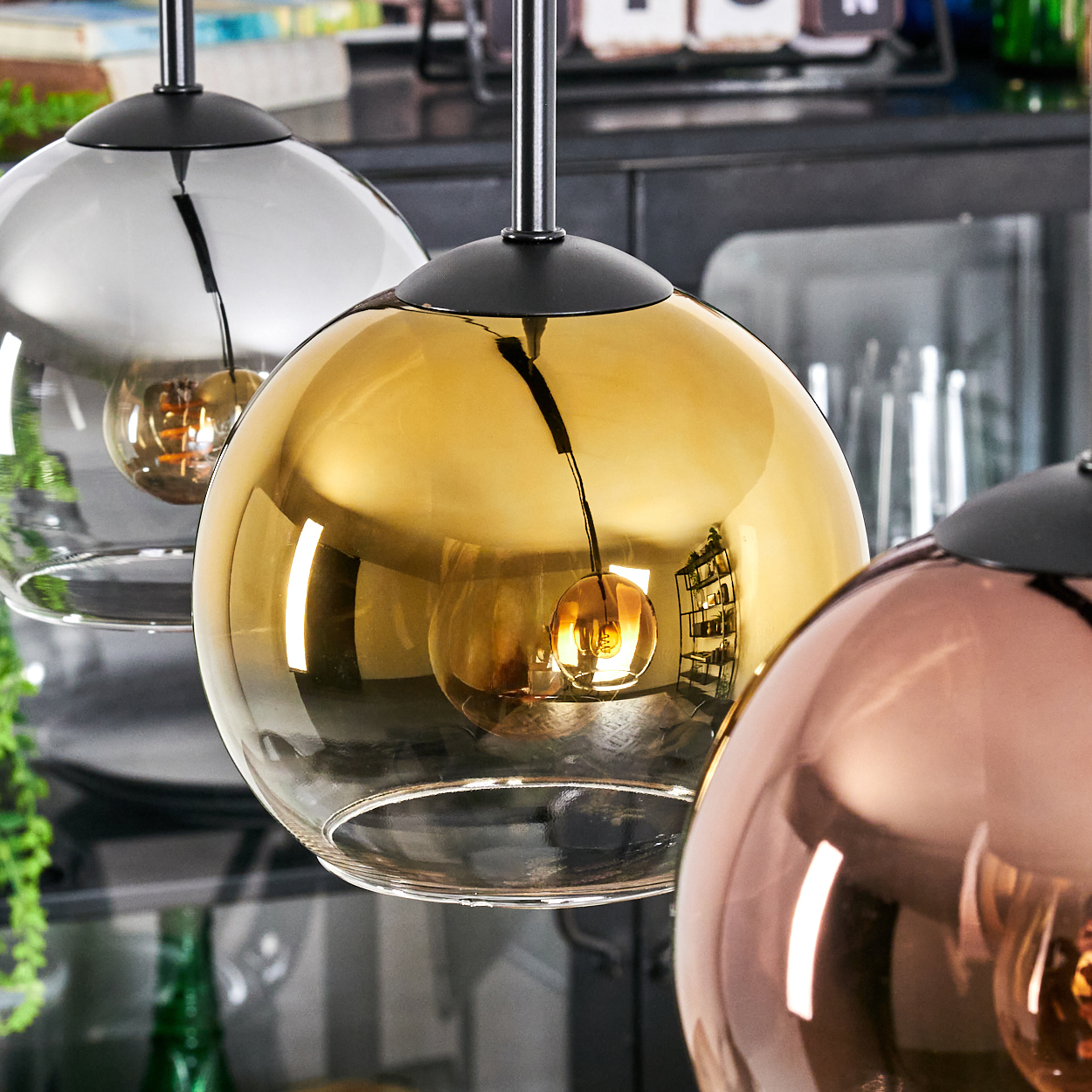 Drei kugelförmige Pendelleuchten, Glas in Silber-, Gold- und Kupferfarben. Jede hat eine schwarze Kappe und ein Kabel. Der Hintergrund ist unscharf.
