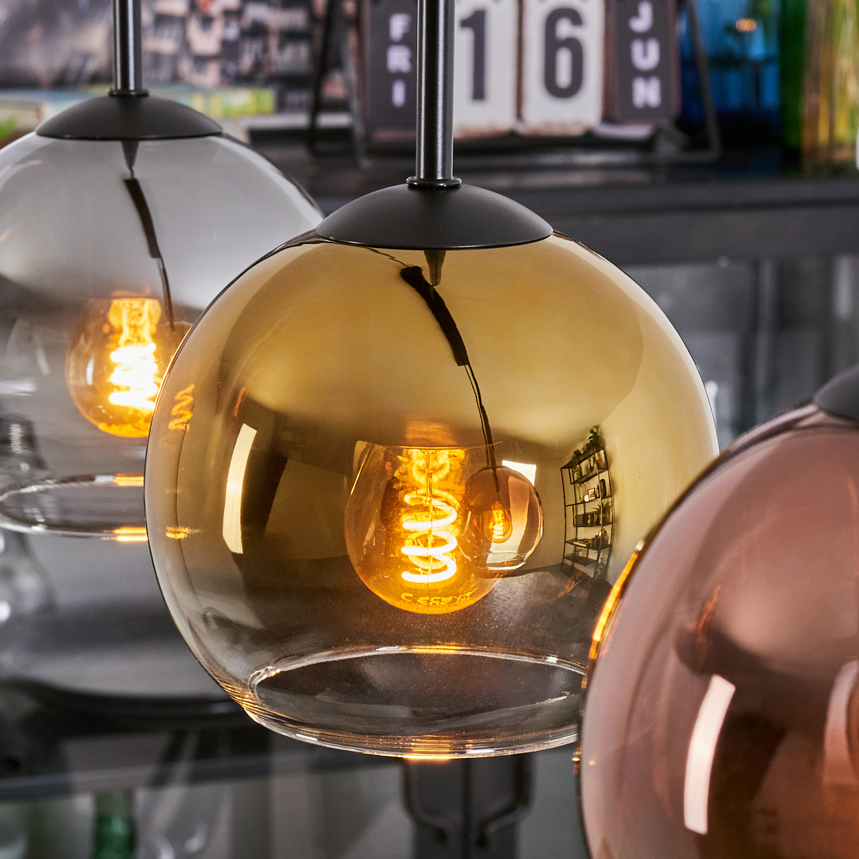 Drei kugelförmige Pendelleuchten mit sichtbaren Filamenten. Das Glas ist goldfarben, grau und kupferfarben getönt. Im Hintergrund ist ein Kalender.