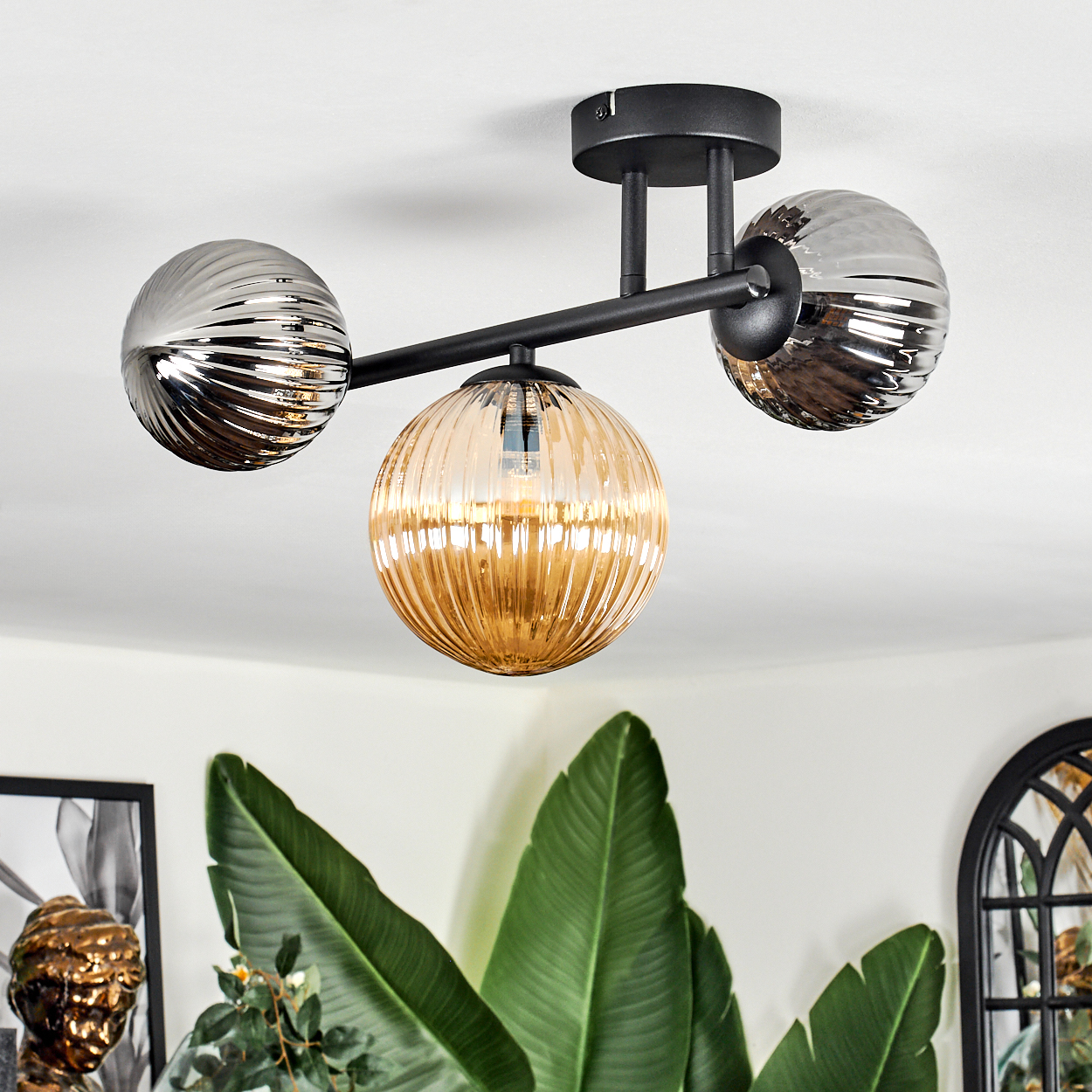 Moderne Deckenlampe mit drei Kugelschirmen, zwei silbernen und einem bernsteinfarbenen. Grüne Blätter und eine dekorative Büste im Hintergrund.