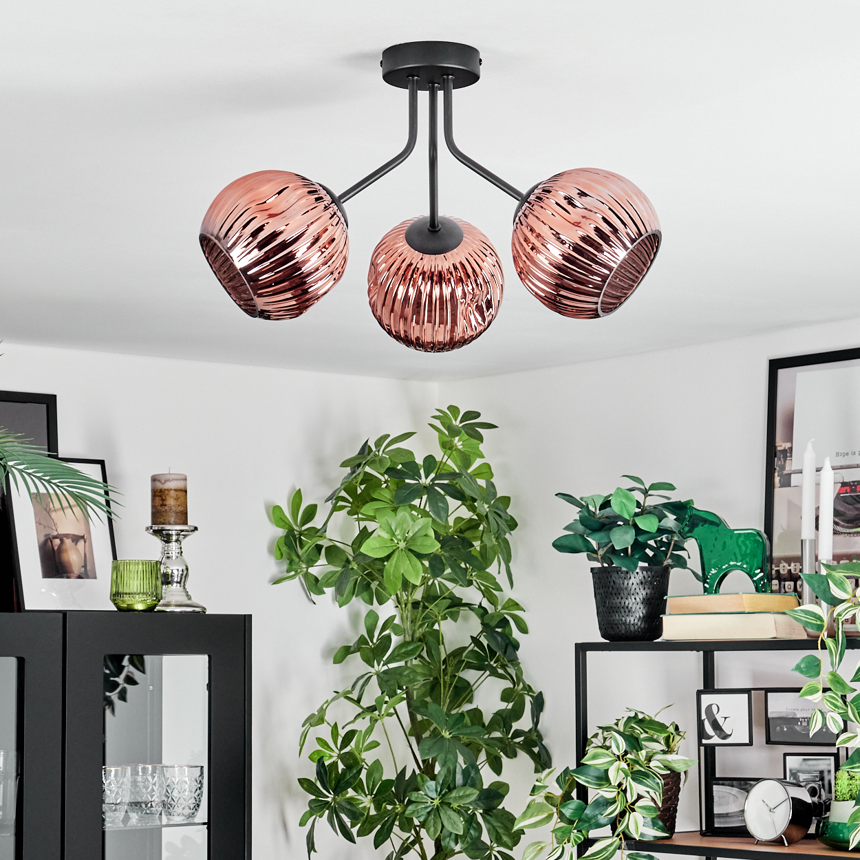 Eine moderne Deckenlampe mit drei kupferfarbenen Glasschirmen. Pflanzen und Dekoration umgeben sie. Schwarze Regale und Schränke sind sichtbar.