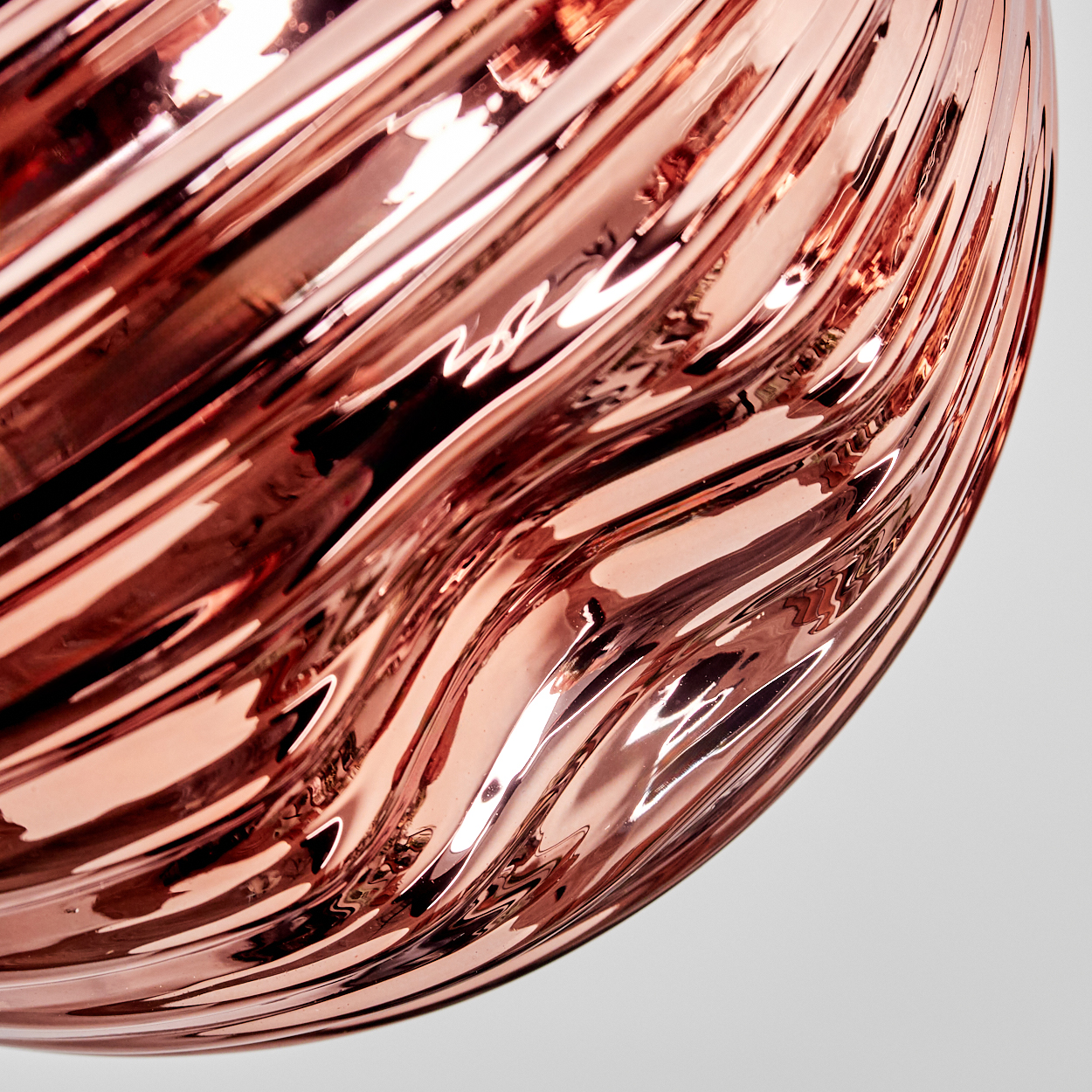 Nahaufnahme einer bronzefarbenen, wellenförmigen, reflektierenden Oberfläche vor weißem Hintergrund. Das Licht reflektiert, wodurch Glanzlichter und Schatten entstehen.