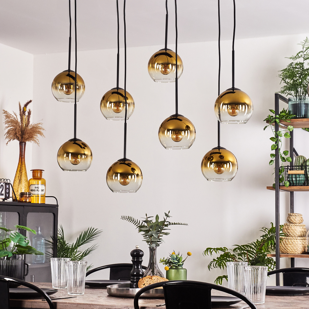 Esszimmer mit acht goldenen Pendelleuchten. Tisch mit Gläsern und Pflanzen. Regale rechts mit Pflanzen und Dekorationen.