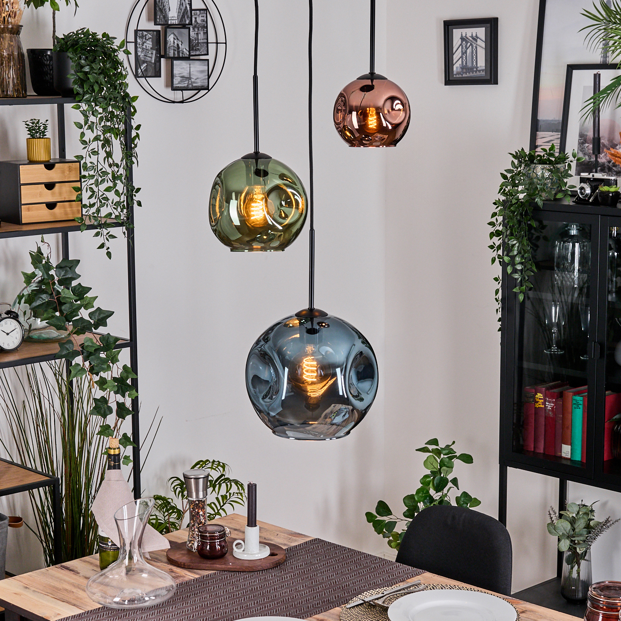 Drei Pendelleuchten mit Glaskugeln hängen über einem Holztisch. Pflanzen und Dekorationen füllen den Hintergrund. Esszimmer-Setting.
