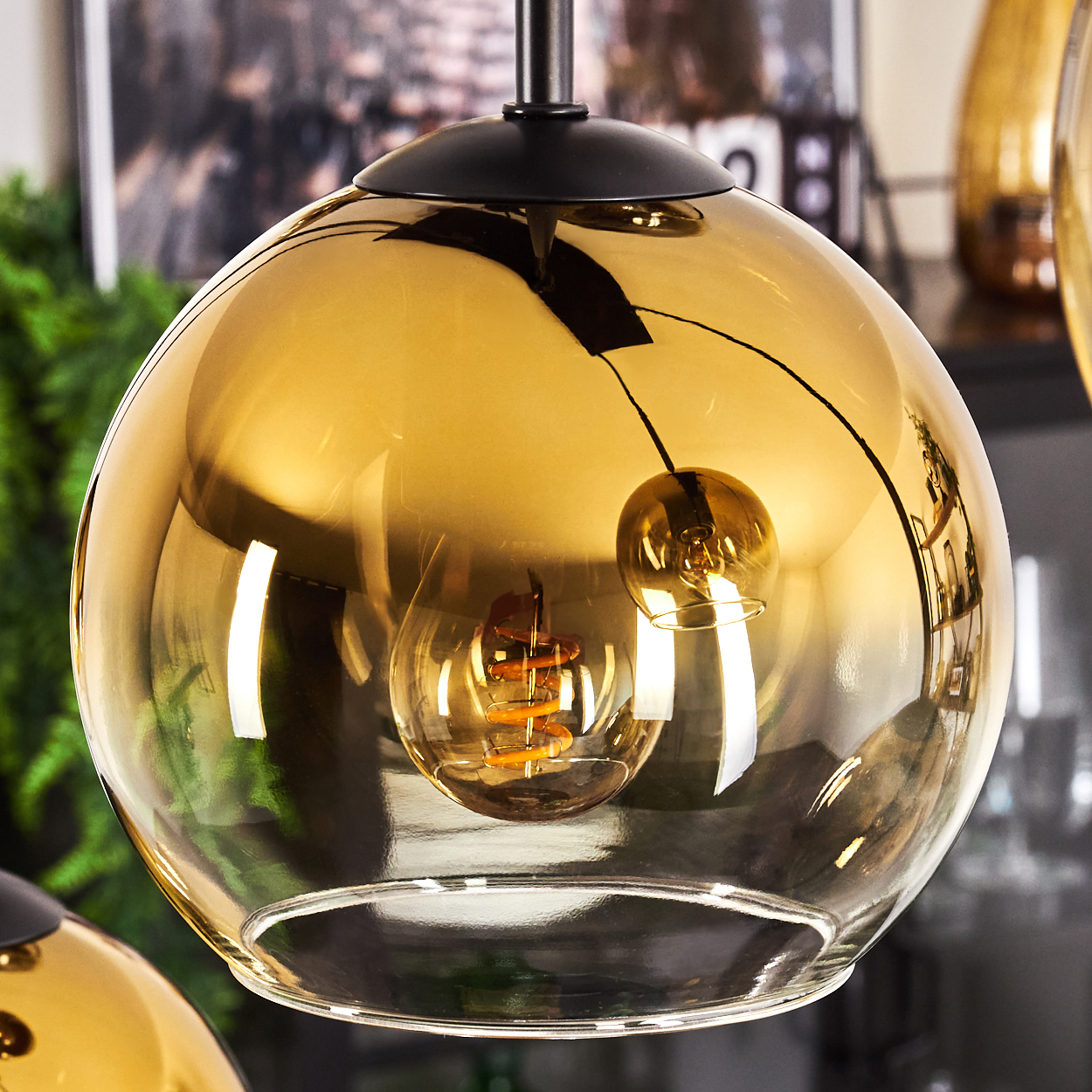 Nahaufnahme einer goldfarbenen Glaslampe mit sichtbarer Glühbirne. Der Hintergrund zeigt verschwommene Dekoration und Grün.