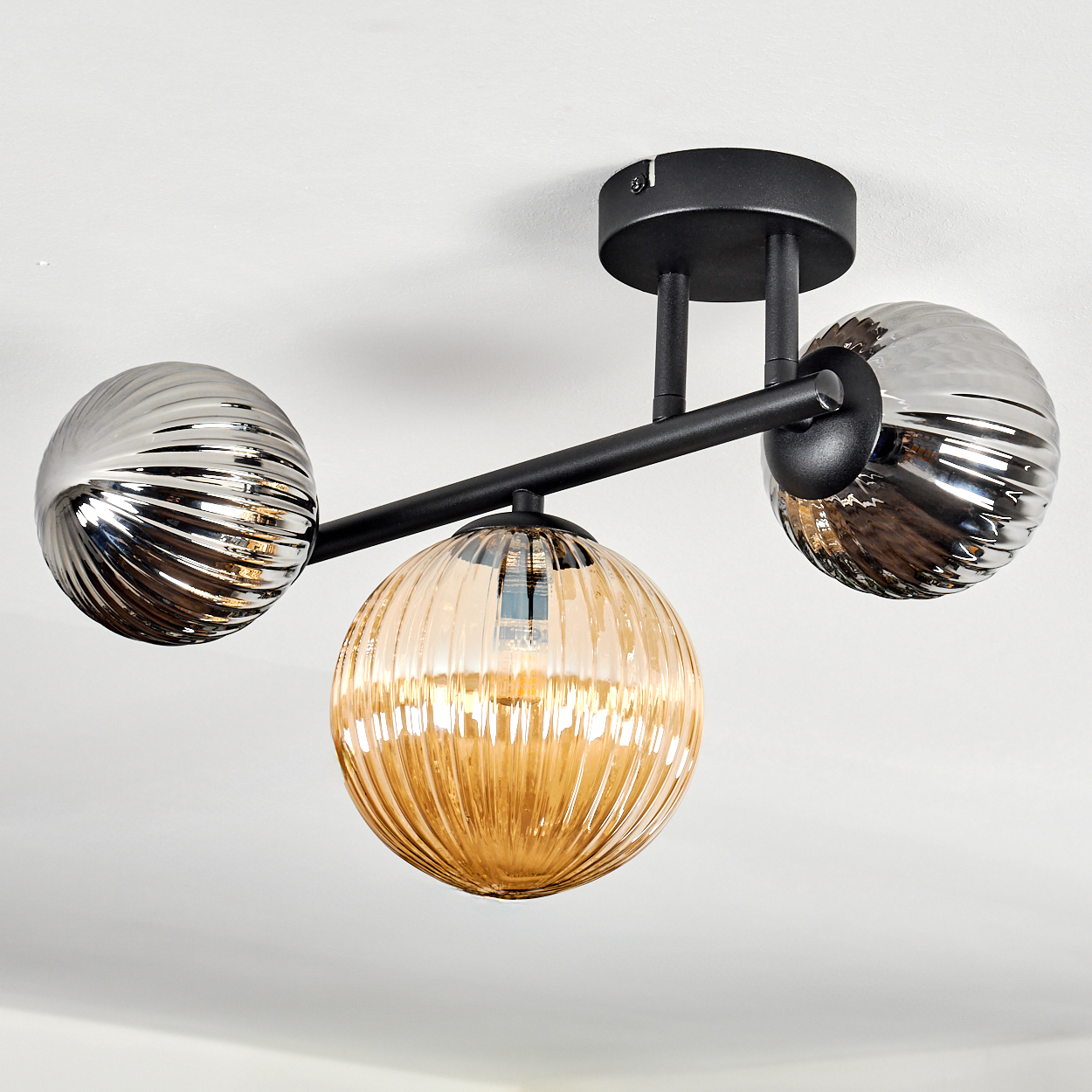 Eine moderne Deckenleuchte mit drei kugelförmigen Lampen. Zwei sind silbern, eine bernsteinfarben. Die Halterung ist schwarz, an einer weißen Decke befestigt.